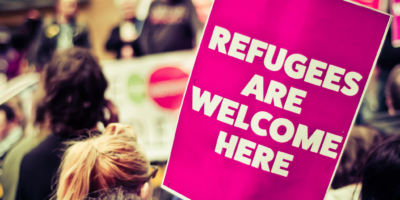 crowd at a protest, person holding sign that says, "refugees are welcome here"