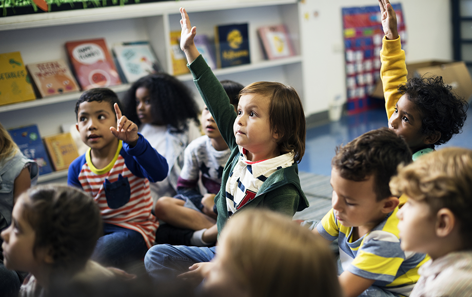 Young elementary students sitting on floor of classroom, several raising their hands